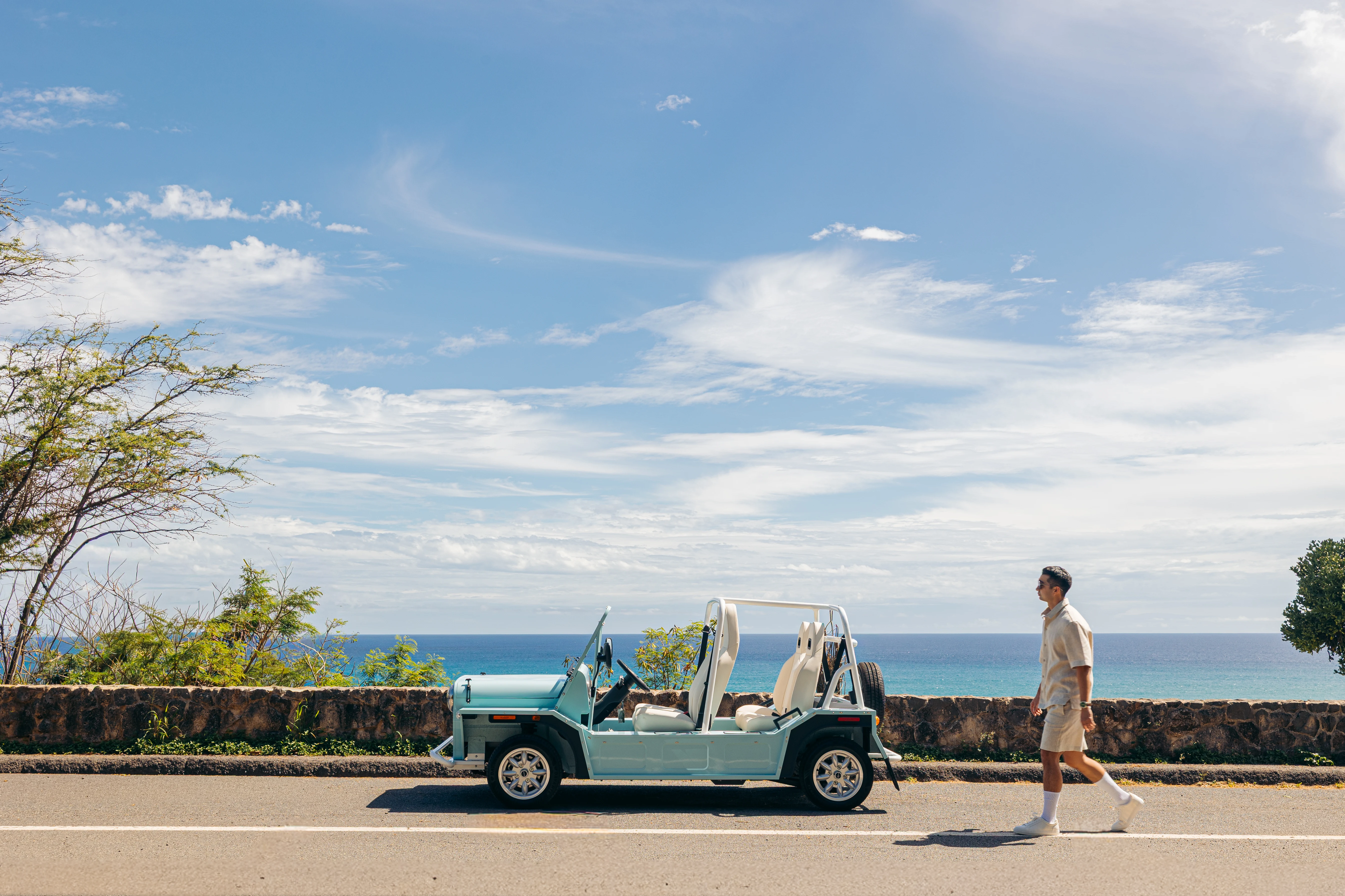 A guest walking toward a seafoam Moke parked on the coast road above the ocean